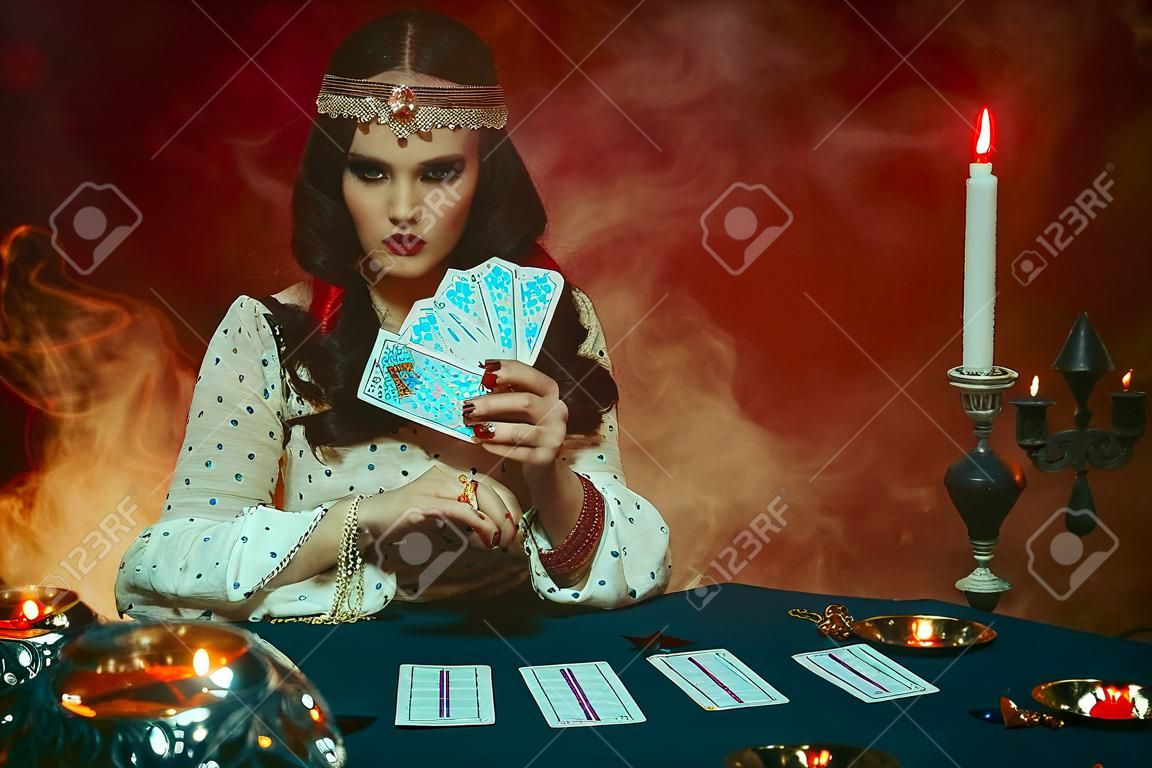 Fantasia bela menina magnífica na imagem de um cigano senta-se à mesa na sala gótica escura. Fato vermelho, roupas vintage, arte preto compõem. Mulher da Fortune teller lendo futuro em cartas de tarô mágico.