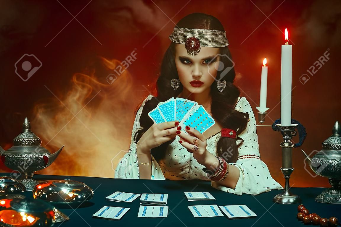 Fantasieschönes prächtiges Mädchen im Bild einer Zigeunerin sitzt am Tisch im dunklen gotischen Raum. Rotes Kostüm, Vintage-Kleidung, schwarzes Kunst-Make-up. Wahrsagerin liest Zukunft auf magischen Tarotkarten.