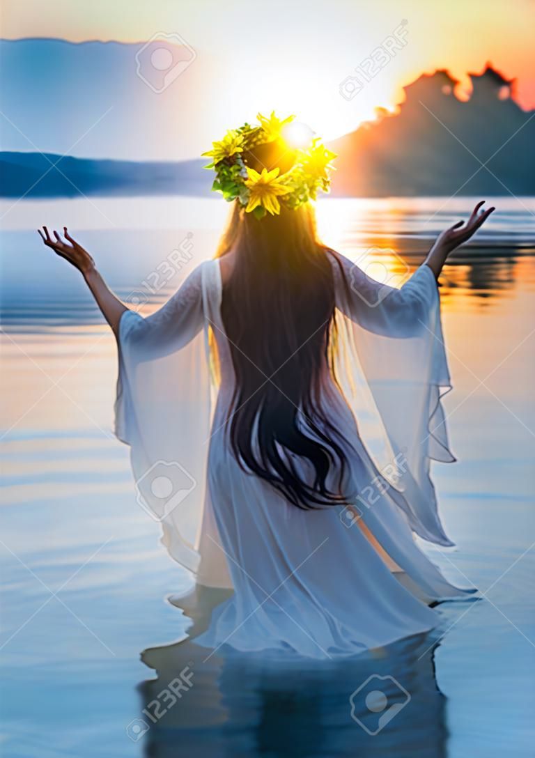 Mulher de fantasia em pé na água mãos levantadas para o céu, orando ao sol. Menina eslovena em grinalda de ervas na cabeça, vestido branco de cabelo longo, feriado pagão de Ivan Kupala. Natureza azul rio pôr do sol. Vista traseira.