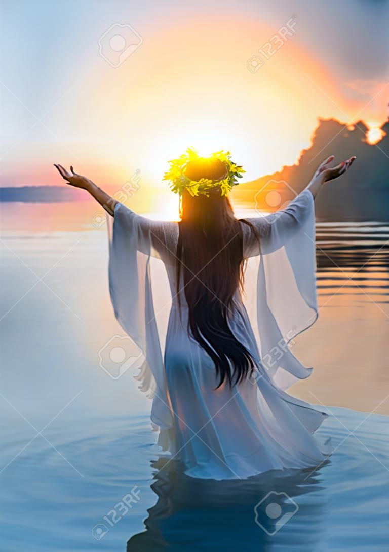 Fantasy woman standing in water hands raised to sky, praying to sun. Slovenian girl in herbal wreath on head, long hair white dress, pagan holiday of Ivan Kupala. Nature blue river sunset. Back view.