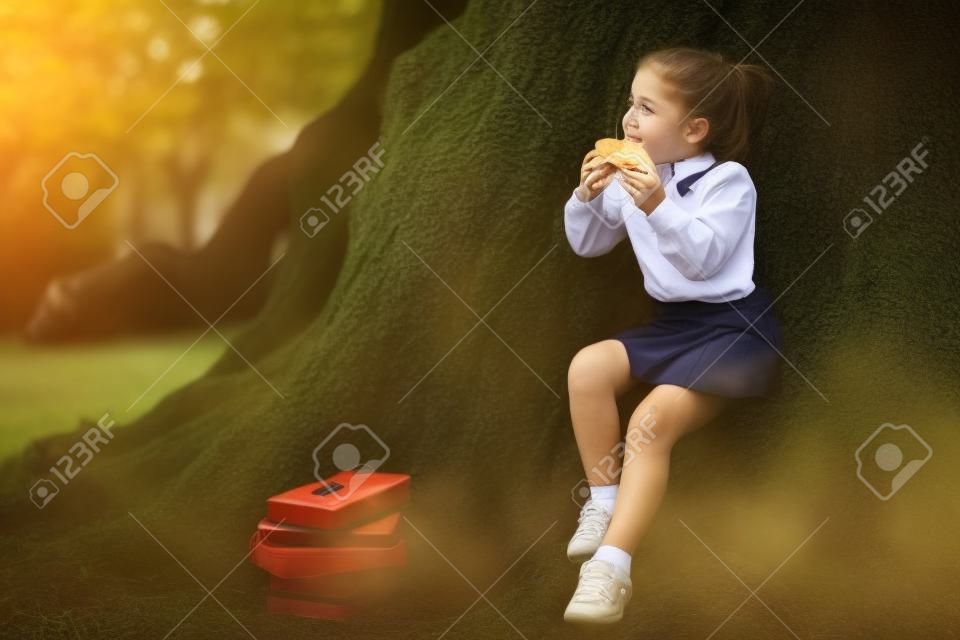 a girl in school uniform, eating a sandwich near a large tree, during a break between classes at school, September 1