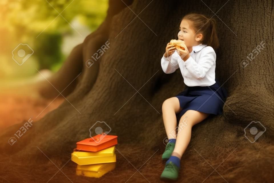 a girl in school uniform, eating a sandwich near a large tree, during a break between classes at school, September 1
