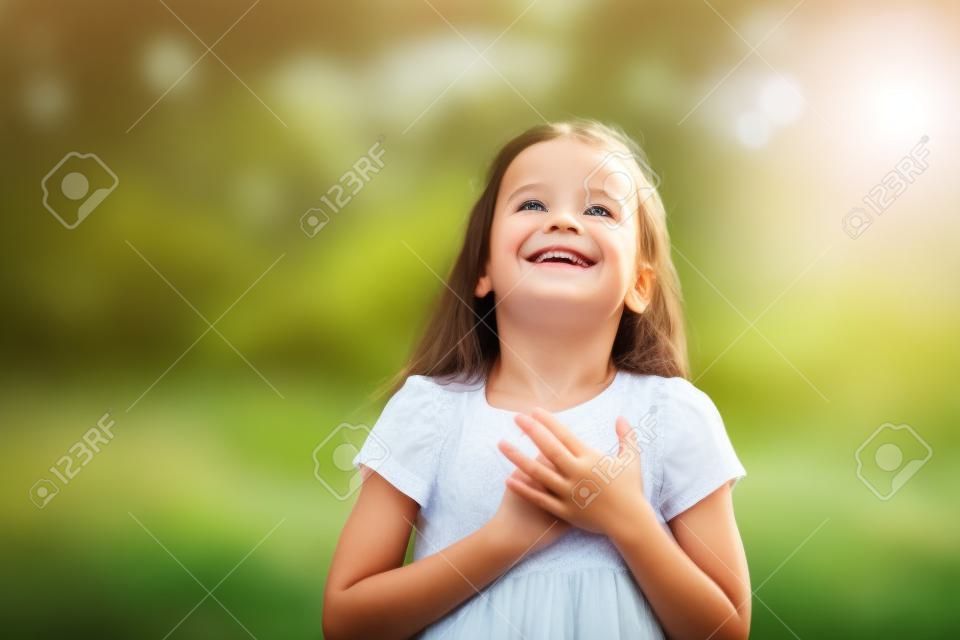 Little girl looking up to at sky with hands on chest, summer nature outdoor. Happy smiling kid feels grateful, wishes dream come true