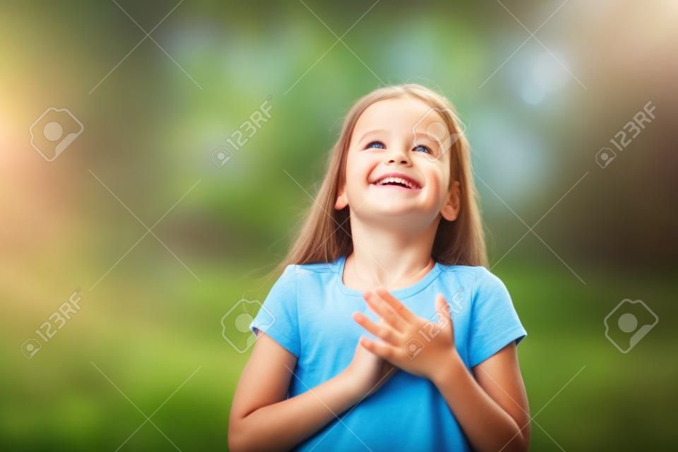 Little girl looking up to at sky with hands on chest, summer nature outdoor. Happy smiling kid feels grateful, wishes dream come true