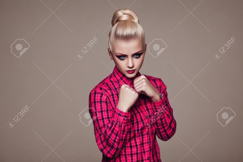 Serious beautiful blonde girl in red, pink checkered shirt, collected bun hairstyle, makeup standing and looking at camera with fists boxing and attack. indoor studio shot. isolated on gray background