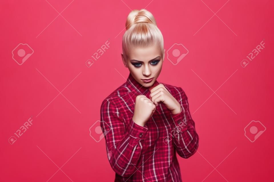 Serious beautiful blonde girl in red, pink checkered shirt, collected bun hairstyle, makeup standing and looking at camera with fists boxing and attack. indoor studio shot. isolated on gray background