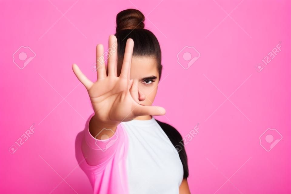 やめる！手のひらで禁止ブロックジェスチャー、否定的な表現を示す上げられた手で立っているカジュアルな服でお団子の髪型を持つ怒っている10代の少女の肖像画。スタジオショット、ピンクの背景