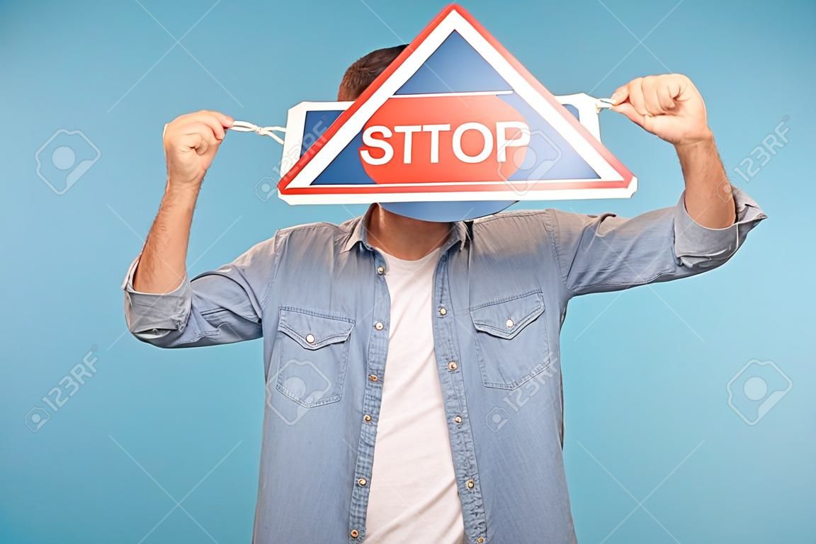 Porträt eines anonymen Mannes im Arbeiter-Jeanshemd, das das Gesicht mit dem Verkehrszeichen Stop als Symbol für Verbote, Beschränkungen und verbotenen Zugang bedeckt. Studioaufnahme im Innenbereich isoliert auf blauem Hintergrund