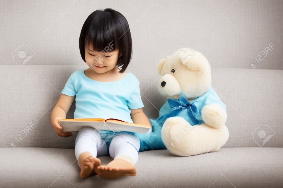Menina chinesa asiática lendo livro com ursinho de pelúcia no sofá na sala de estar.