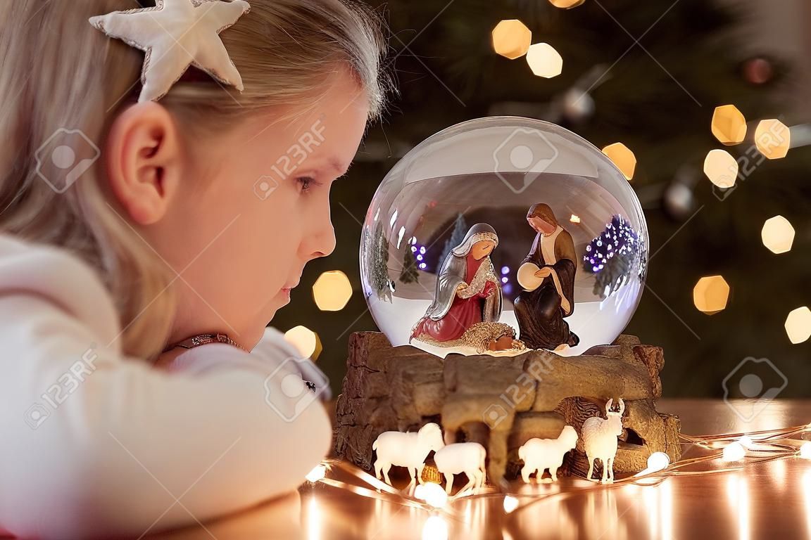 Girl looking at a glass ball with a scene of the nativity of Jesus Christ in a glass ball on a Christmas tree