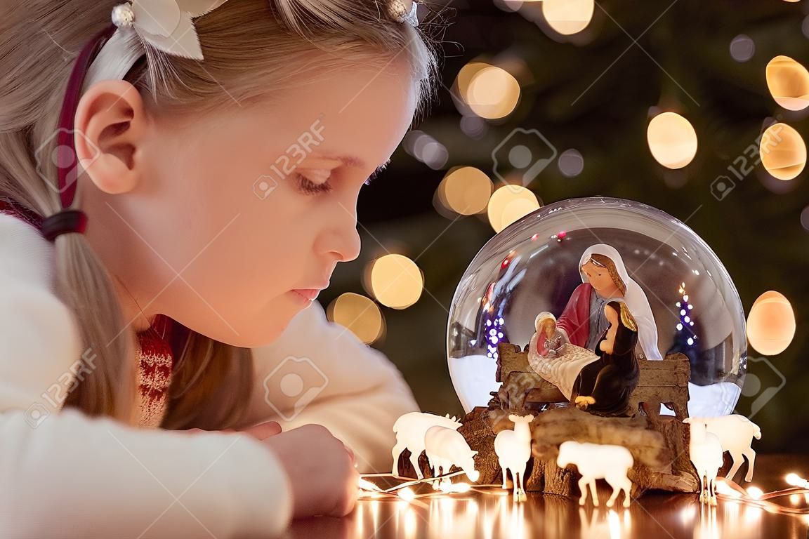 Chica mirando una bola de cristal con una escena de la natividad de Jesucristo en una bola de cristal en un árbol de Navidad