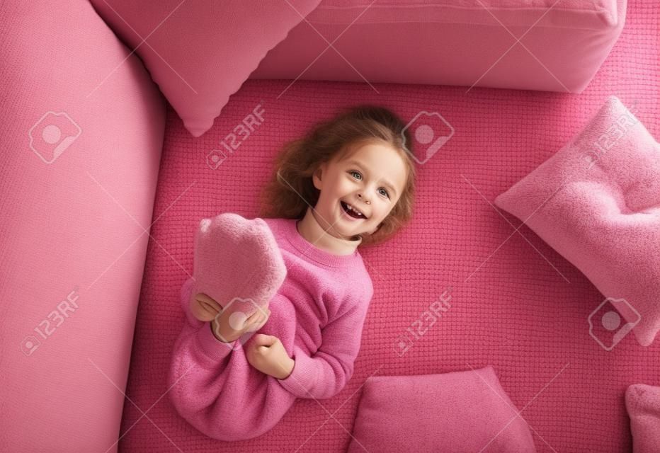 a little girl in a pink sweatshirt is lying on the sofa among the pillows with her legs up. Home games.