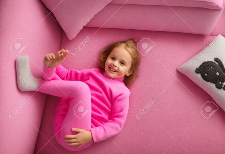 uma menina em um suéter rosa está deitado no sofá entre os travesseiros com as pernas para cima.