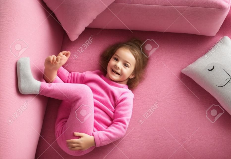 a little girl in a pink sweatshirt is lying on the sofa among the pillows with her legs up. Home games.