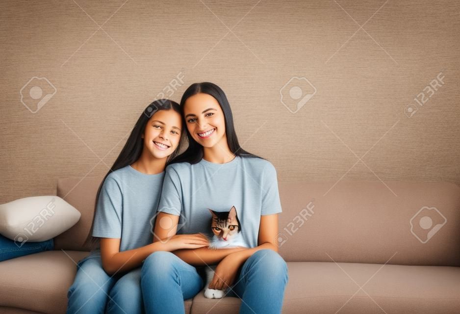Mãe e filha estão sentadas no sofá com os braços em volta uma da outra segurando um gato.