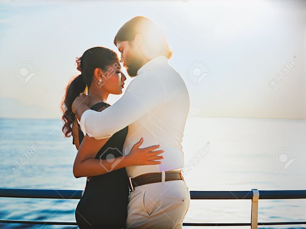 Portrait of young attractive couple dancing latin bachata near sea or ocean. Sunlight background. Man gently hugs his beloved girl and looks at her with admiration