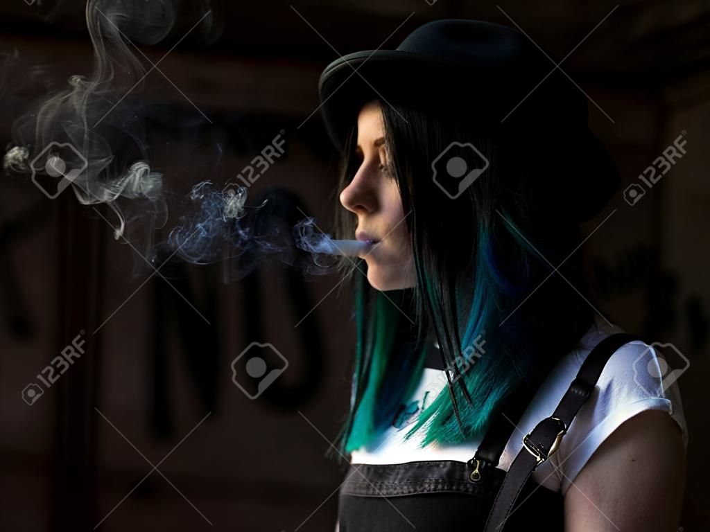 Chica emo fumando cigarrillos. Mujer punk callejera o hipster con cabello teñido de color azul, sombrero, piercing, lentes, túneles de orejas y peinados inusuales se encuentra en el patio trasero. hermoso humo.
