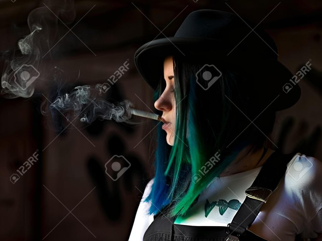 Chica emo fumando cigarrillos. Mujer punk callejera o hipster con cabello teñido de color azul, sombrero, piercing, lentes, túneles de orejas y peinados inusuales se encuentra en el patio trasero. hermoso humo.