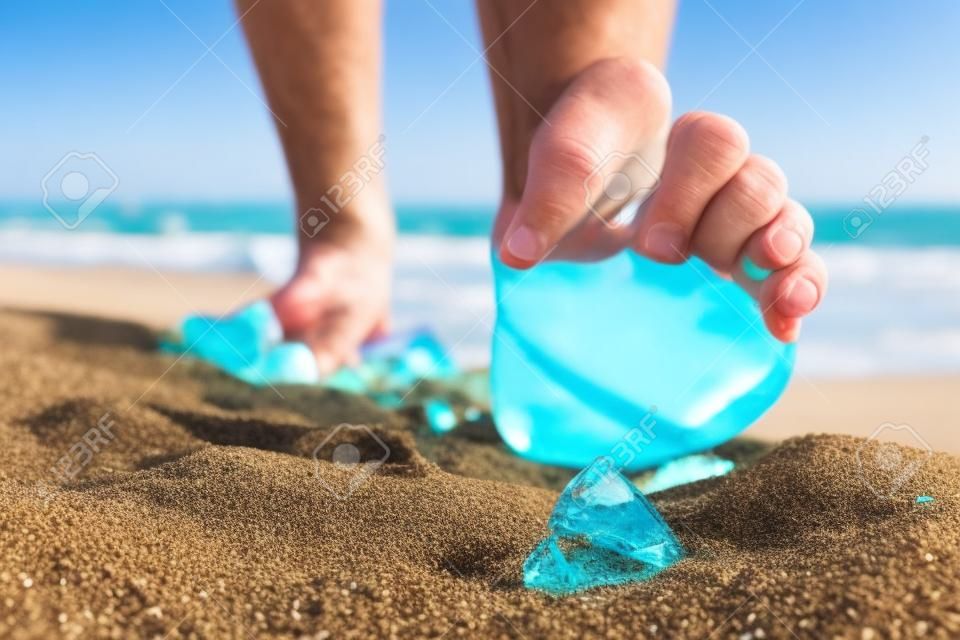 男人去海灘，踩著碎玻璃瓶的碎片一個，這是躺在與惡劣環境的地方散落的沙子上的風險