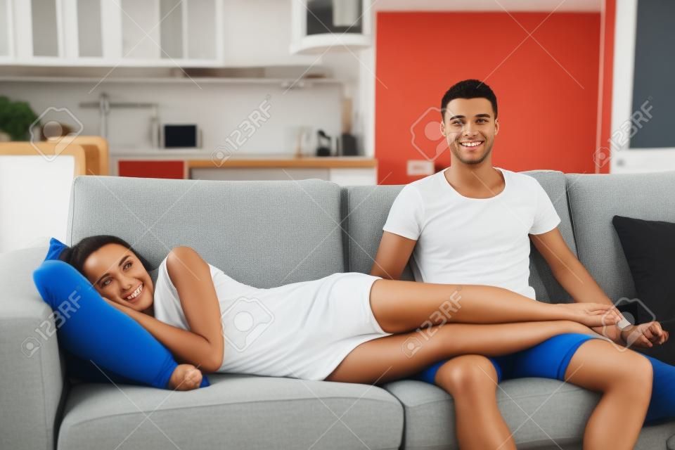 Young Couple Watching Television Together on Sofa - Woman Lying Down Relaxing with Feet Up on Lap of Man