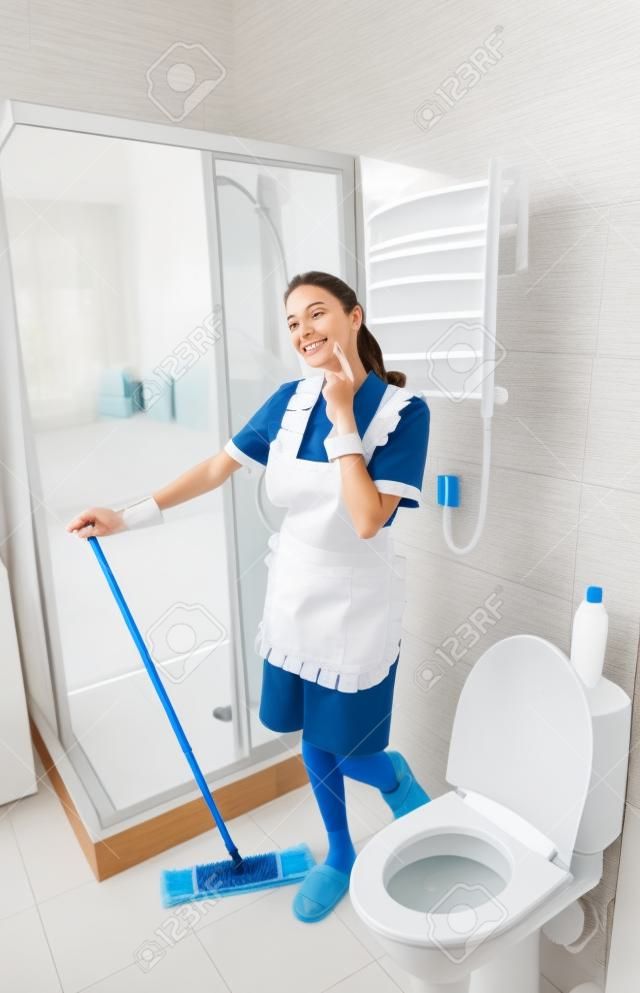 Pretty young housekeeper or maid relaxing on the job standing staring thoughtfully into the air as she cleans the bathroom with a mop