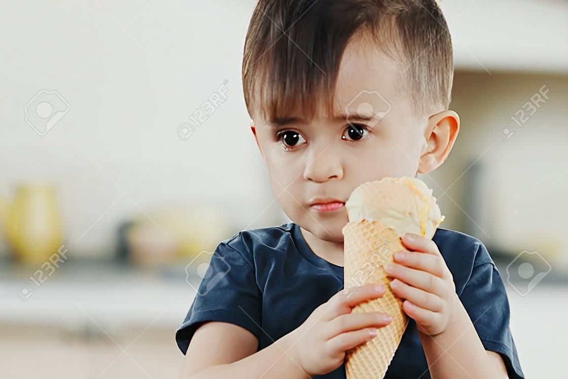 A child in a dark-blue t-shirt in the bright kitchen eating a waffle ice cream cone in the summer house