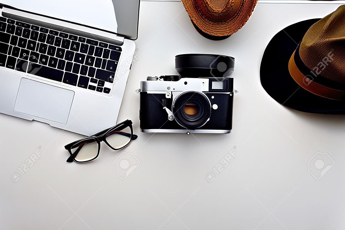 Top view of laptop, eyeglasses, photo camera, hat and a cup of coffee on white surface