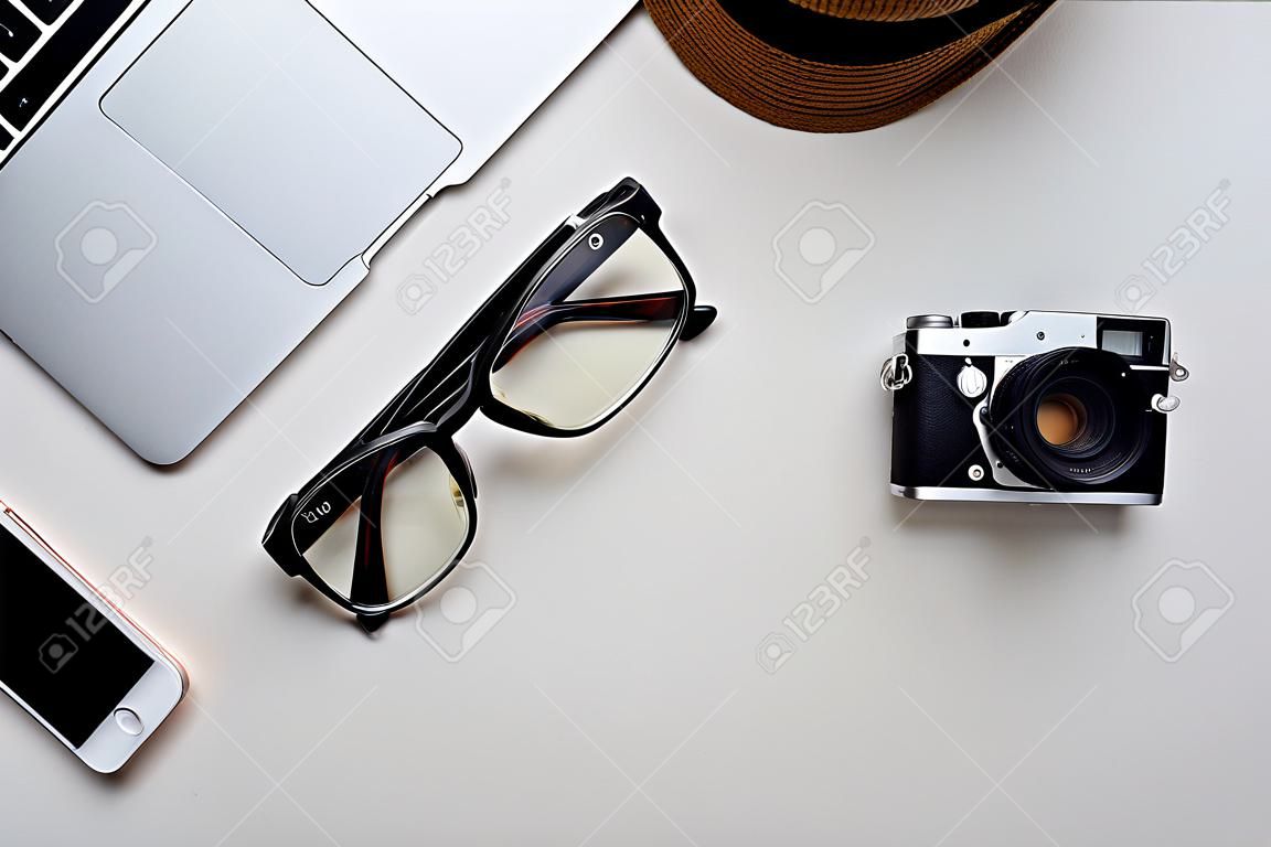 Widok z góry laptopa, okularów, aparatu fotograficznego, kapelusza i filiżanki kawy na białej powierzchni