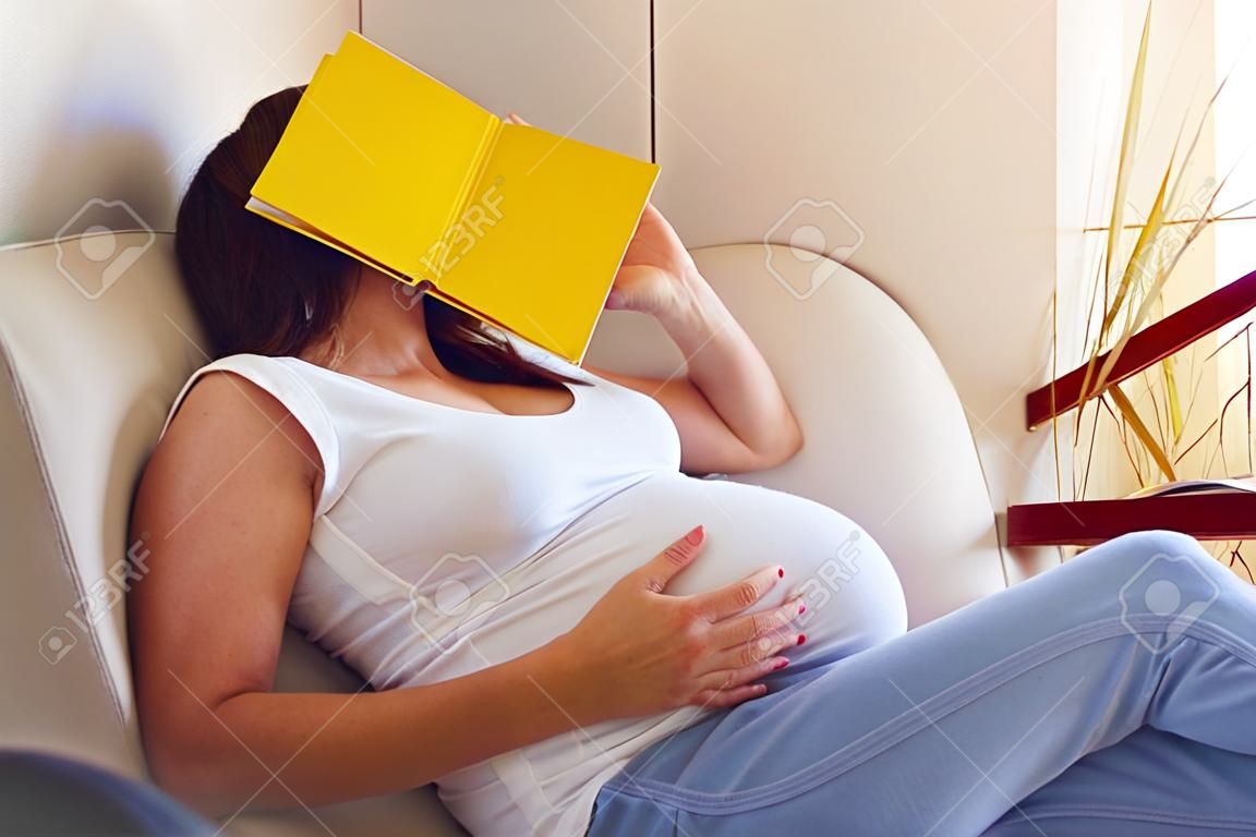 Seitenansicht der schwangeren Frau müde vom Lesen, schlafend auf Sofa mit einem Buch auf ihrem Gesicht
