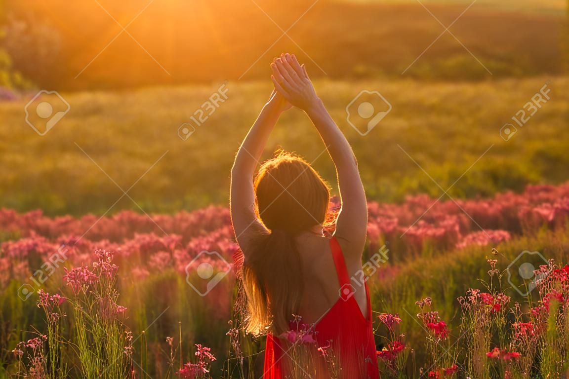 Girl in red dress staying by back with hands up  among blooming Sally field in sunset