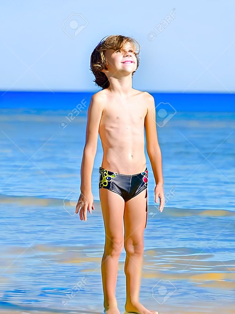 Lustiges Kind in schwarzen Badehosen in einer Meereskappe mit Blick in die Ferne auf dem Hintergrund des blauen Meeres und des Himmels