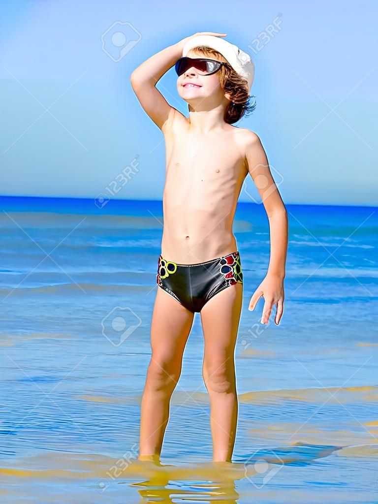 Bambino divertente in costume da bagno nero in un berretto marino che guarda in lontananza sullo sfondo del mare e del cielo blu blue