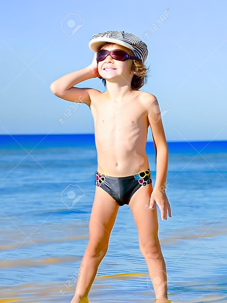 Criança engraçada nos troncos de natação pretos em uma tampa do mar que olha na distância no fundo do mar azul e do céu