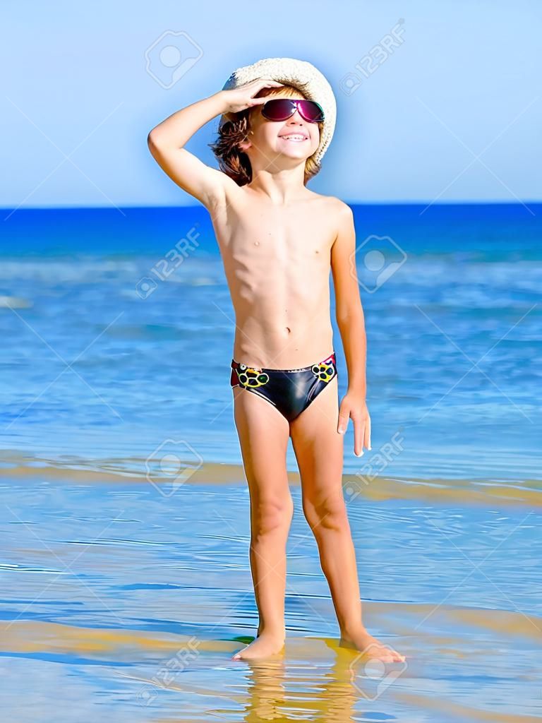 Zabawne dziecko w czarnych kąpielówkach w morskiej czapce patrzące w dal na tle błękitnego morza i nieba