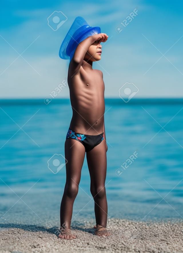 Enfant drôle dans des maillots de bain noirs dans un chapeau de mer regardant au loin sur le fond bleu de la mer et du ciel