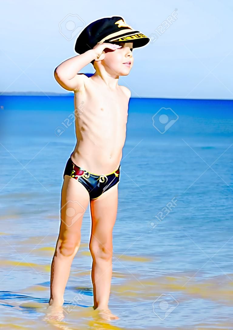 Enfant drôle dans des maillots de bain noirs dans un chapeau de mer regardant au loin sur le fond bleu de la mer et du ciel