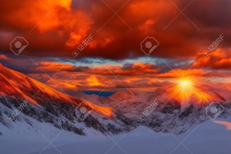 mountain peak sunset landscape with gloomy dramatic mainly cloudy sky and orange and red sun beams at snow