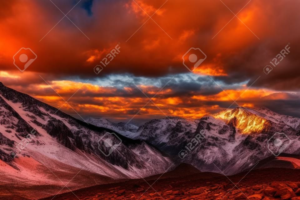 mountain peak sunset landscape with gloomy dramatic mainly cloudy sky and orange and red sun beams at snow