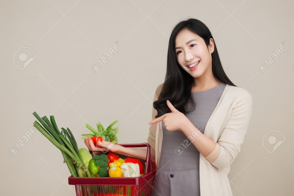 野菜や食料品でいっぱいの買い物かごを持つ美しいアジアの女性、白い背景に隔離されたスタジオショット