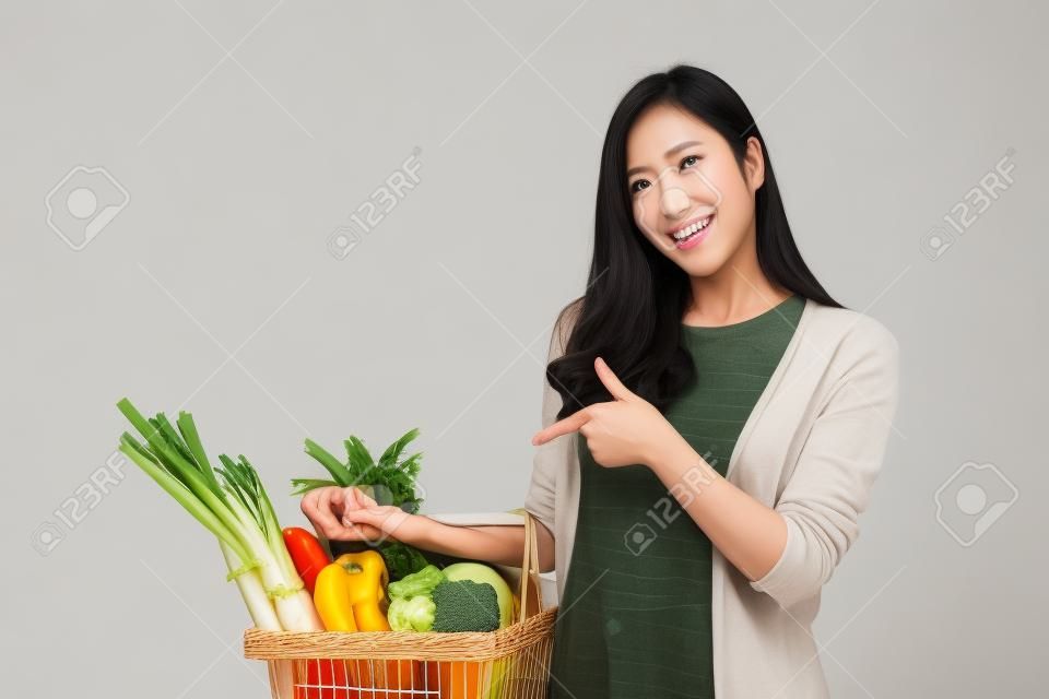 Beautiful Asian woman holding shopping basket full of vegetables and groceries, studio shot isolated on white background