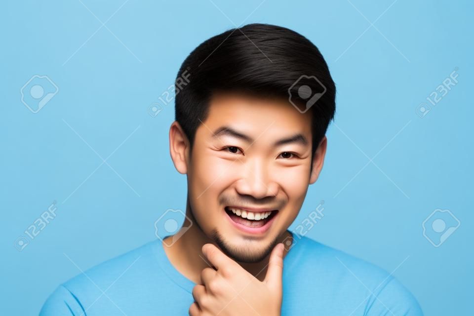 Happy smiling young handsome Asian man face with hand touching chin studio shot isolated on light blue background