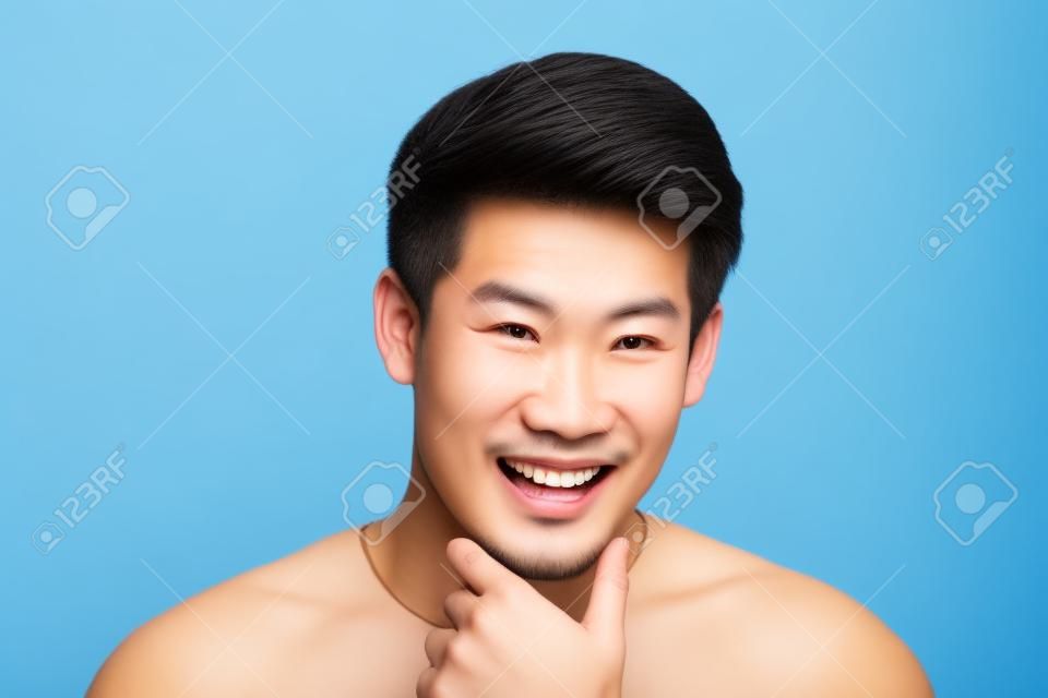 Felice sorridente giovane uomo asiatico bello faccia con la mano che tocca il mento studio shot isolato su sfondo azzurro