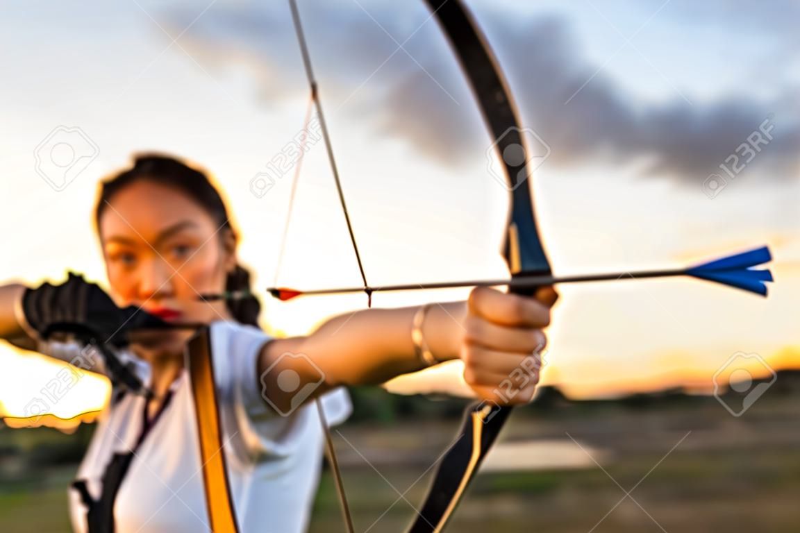 young female archer, archery, shoot arow with bow in nature field to target, success concept, at field for sport exercise at sunset time