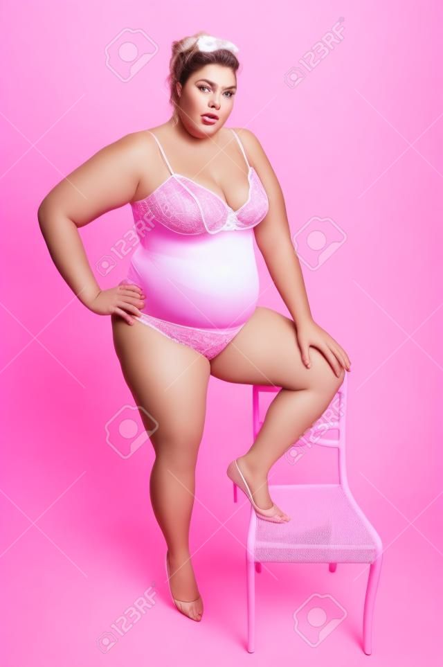 Eine große übergewichtigen Frau in pink Lingerie stehen, mit einem Bein auf einem Stuhl, im Studio, white background.