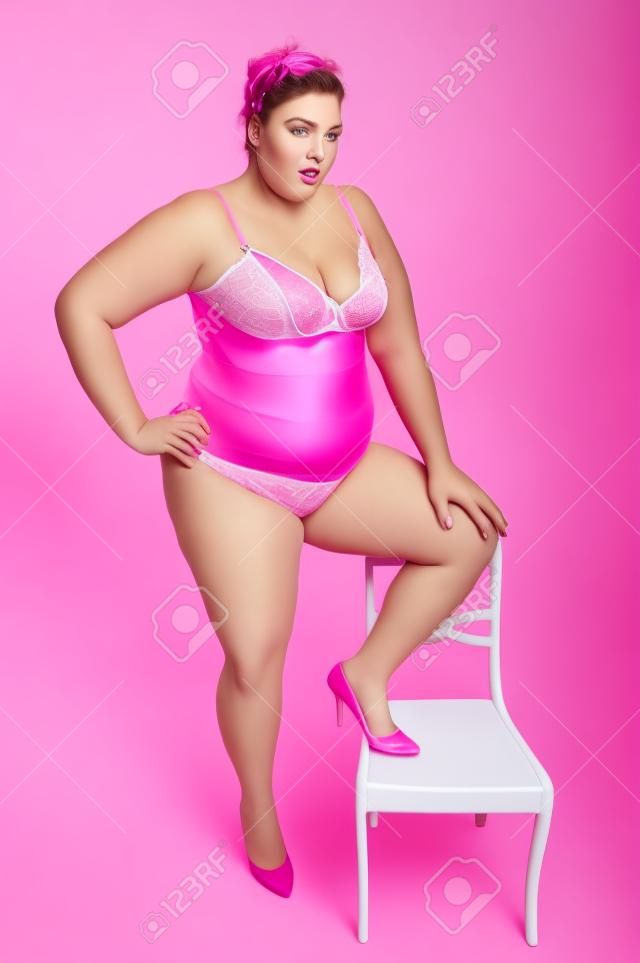 一個大女人超重粉紅色內衣站立，把一條腿的椅子上，在錄音室，為白色背景。