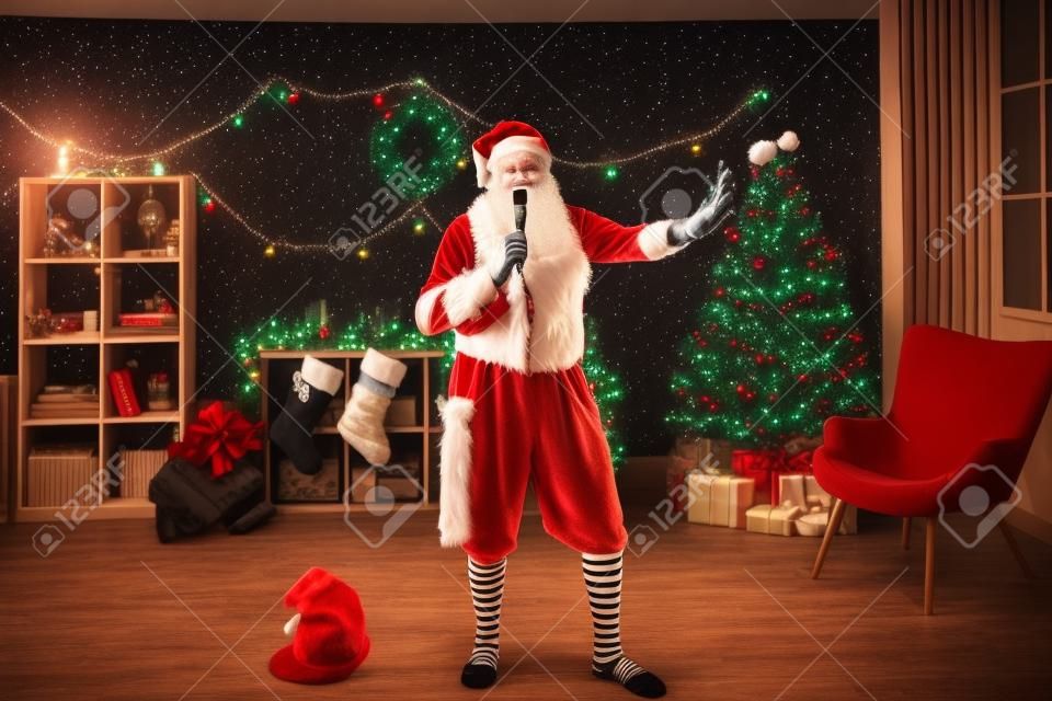 Grappige opa in onderhemd, Santa Claus baard, hoed en gestreepte sokken met Mike en zingen Kerstliedjes op karaoke nacht feest thuis. Senior man hebben plezier en ontspannen op nieuwjaarsvakantie