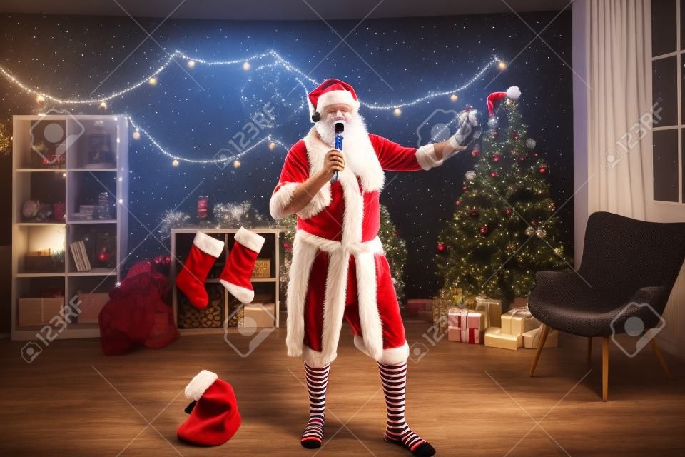 Vovô engraçado em undershirt, barba de Papai Noel, chapéu e meias listradas segurando mike e cantando canções de Natal na festa de noite de karaoke em casa.