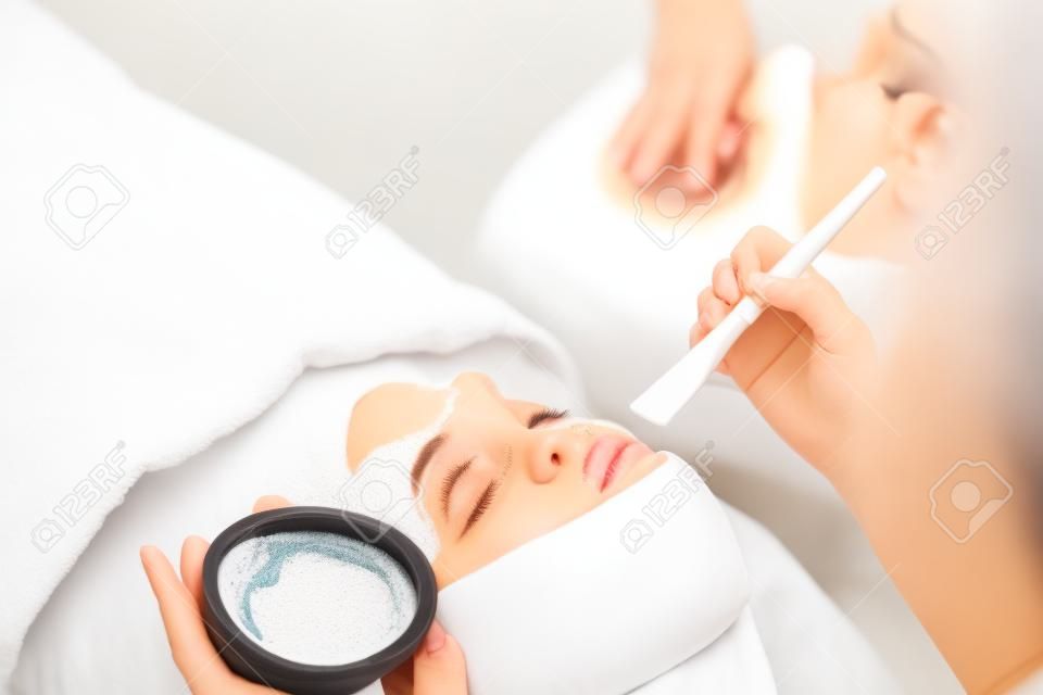 Mãos de cosmetologista colocando máscara cremosa especial para limpeza facial para mulher relaxante no salão de beleza, vista traseira. Tratamento facial, massagem, cuidados com a pele, conceito de cosmetologia