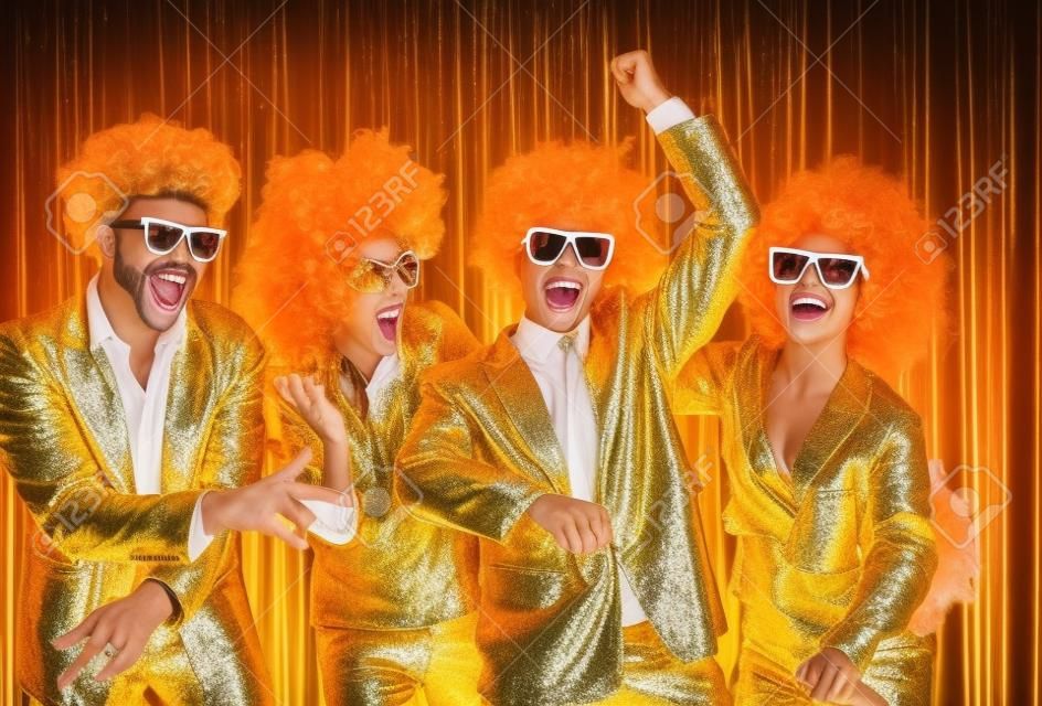 Happy people dancing   style on stage with shiny golden background. Group of friends disguised in boas, glasses, sequin jackets and funny silly curly wigs having fun at night club disco party