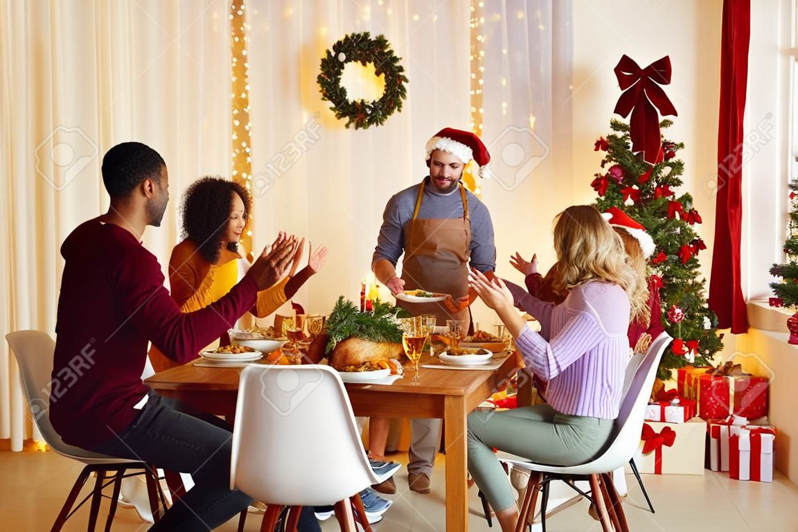 Family and friends dining at home celebrating Christmas eve with traditional food and decoration. Multiracial people sit at table in living room and applaud their male friend who serves delicious meal